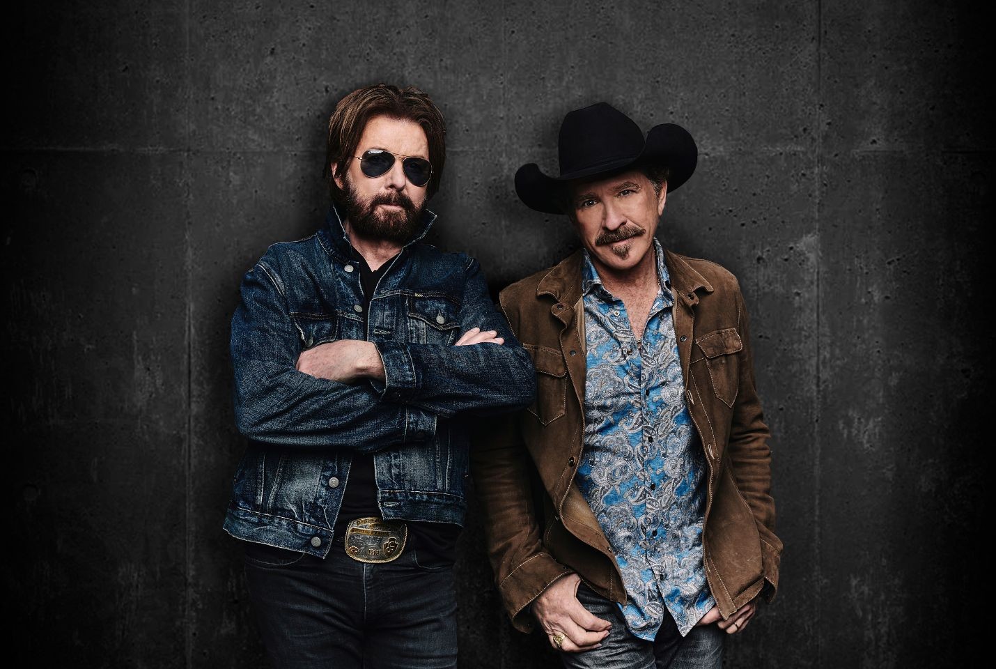 Brooks & Dunn stood against a black wall one wearing a denim jacket and black sunglasses, the other a brown jacket and black cowboy hat